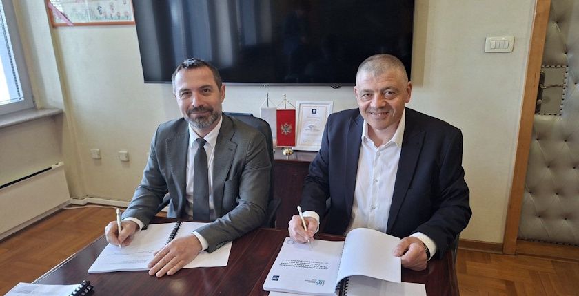Two men in suits signing official building contract documents in an office.